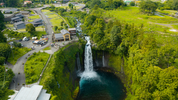 Aerial view of Shiraito Falls cascading into a deep blue pool surrounded by lush green forest and a nearby building with a parking area in Japan. Aerial view of Shiraito Falls cascading into a deep blue pool surrounded by lush green forest and a nearby building with a parking area in Japan.