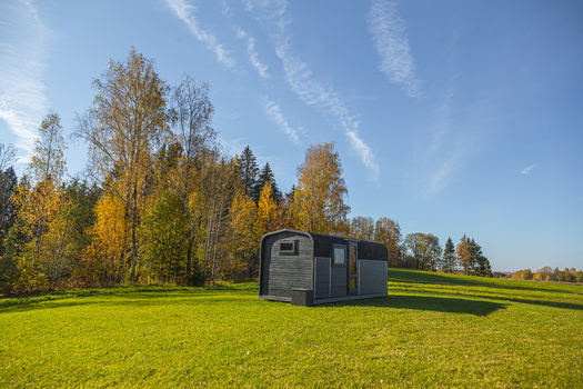 View of a modern tiny house cabin situated in a grassy field with autumn foliage and forest in the background under a clear blue sky Latvia. View of a modern tiny house cabin situated in a grassy field with autumn foliage and forest in the background under a clear blue sky Latvia.