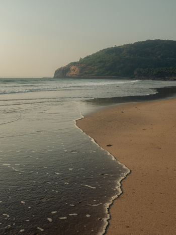View of the serene shoreline kissed by gentle waves under a hazy sky with a tree covered hill rising in the distance Chiplun Maharashtra India. View of the serene shoreline kissed by gentle waves under a hazy sky with a tree covered hill rising in the distance Chiplun Maharashtra India.