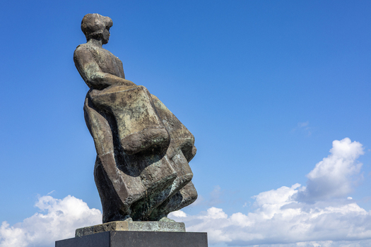 View of the Fisherman s Monument featuring a bronze statue of a woman looking out to sea under a clear blue sky with white clouds in Urk Flevoland Netherlands. View of the Fisherman s Monument featuring a bronze statue of a woman looking out to sea under a clear blue sky with white clouds in Urk Flevoland Netherlands.