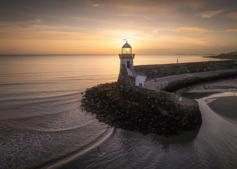 Aerial view of the lighthouse at Balbriggan Harbour illuminated by the warm glow of sunset with calm waters and the stone pier extending into the sea in Balbriggan County Dublin Ireland. Aerial view of the lighthouse at Balbriggan Harbour illuminated by the warm glow of sunset with calm waters and the stone pier extending into the sea in Balbriggan County Dublin Ireland.