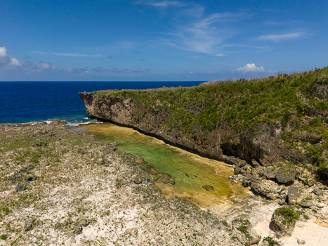 Aerial view of Chigit Beach featuring rugged limestone cliffs turquoise coastal waters and rocky terrain under a clear blue sky in Northern Tinian Tinian Northern Mariana Islands. Aerial view of Chigit Beach featuring rugged limestone cliffs turquoise coastal waters and rocky terrain under a clear blue sky in Northern Tinian Tinian Northern Mariana Islands.