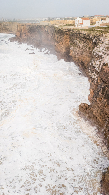 Aerial view of the historic Sagres Fortress perched on steep rocky cliffs with powerful white waves crashing against the shore under a misty sky in Sagres Faro Portugal. Aerial view of the historic Sagres Fortress perched on steep rocky cliffs with powerful white waves crashing against the shore under a misty sky in Sagres Faro Portugal.