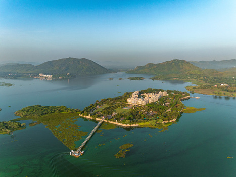 Aerial view of Taj Lake Palace on an island in Lake Pichola surrounded by green hills and calm water with a stone walkway connecting it to the shore in Udaipur Rajasthan India. Aerial view of Taj Lake Palace on an island in Lake Pichola surrounded by green hills and calm water with a stone walkway connecting it to the shore in Udaipur Rajasthan India.