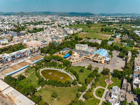 Aerial view of the Udaipur City Railway Station area featuring a large station building a landscaped park with an oval pond and surrounding urban streets in Udaipur Rajasthan India.