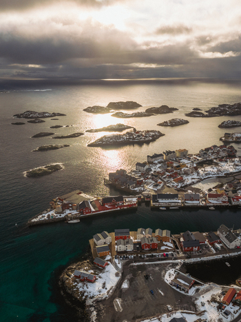 Aerial view of sunlight piercing through dramatic clouds over Henningsv&aelig;r s vibrant red buildings nestled along the rugged coastline Nordland Norway.