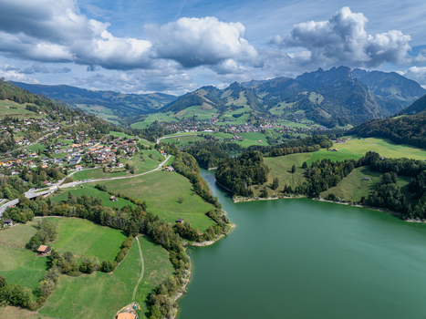 Aerial view of Lac de Montsalvens surrounded by lush green hills small villages and the Swiss Alps under a blue sky with clouds in Gruyere District Fribourg Switzerland. Aerial view of Lac de Montsalvens surrounded by lush green hills small villages and the Swiss Alps under a blue sky with clouds in Gruyere District Fribourg Switzerland.