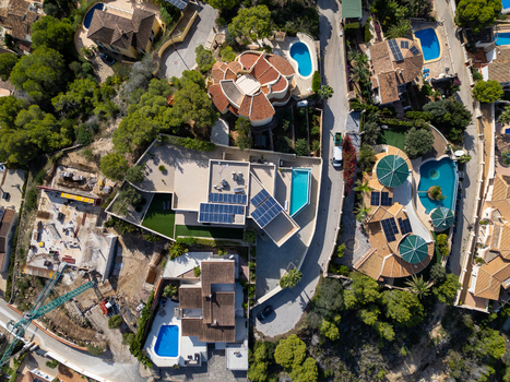 Aerial view of residential villas with swimming pools solar panels and a construction site surrounded by pine trees and winding roads in Moraira Valencian Community Spain.