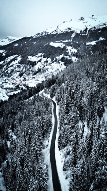 Aerial view of a road winding through a snow laden forest contrasting sharply with the dark asphalt and leading towards the snow capped mountains Grimentz Valais Switzerland.