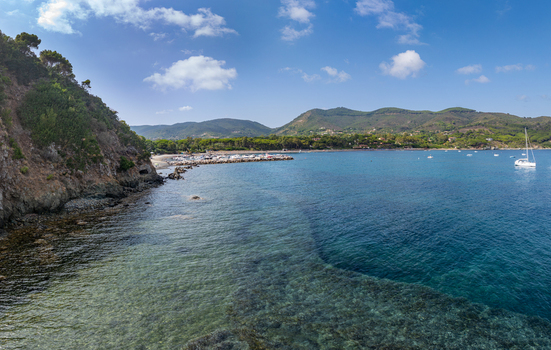 Aerial view of Lacona Beach with its clear turquoise waters rocky coastline and small boats anchored in the bay under a bright blue sky in Lacona Tuscany Italy.