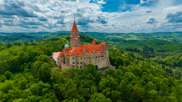Aerial view of Bouzov Castle with its iconic red roofs and tall tower surrounded by dense green forests and rolling hills under a cloudy sky in Bouzov Olomoucky kraj Czechia. Aerial view of Bouzov Castle with its iconic red roofs and tall tower surrounded by dense green forests and rolling hills under a cloudy sky in Bouzov Olomoucky kraj Czechia.