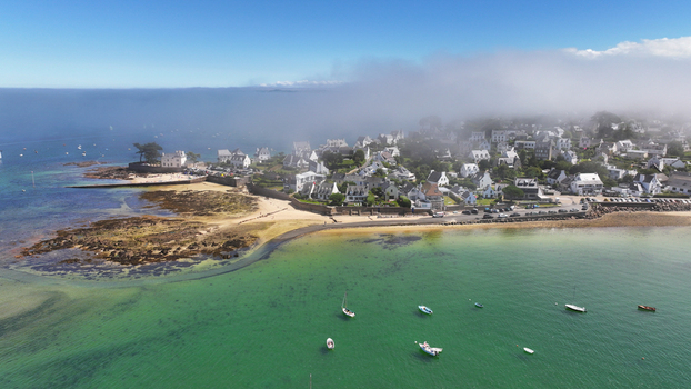 Aerial view of the shoreline where the land meets the sea with boats dotting the clear water and buildings shrouded in mist Carantec Bretagne France.
