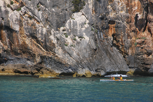 View of two people kayaking in the turquoise waters of the Gulf of Orosei beneath a towering rocky cliff in Italy. View of two people kayaking in the turquoise waters of the Gulf of Orosei beneath a towering rocky cliff in Italy.