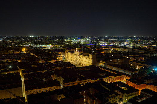 Aerial view of the illuminated Ducal Palace of Modena at night surrounded by the historic city center with glowing streetlights and dark rooftops in Modena Emilia Romagna Italy.