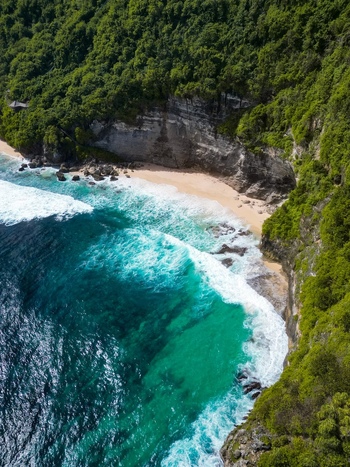 Aerial view of Pantai Nunggalan with turquoise waves crashing against the sandy beach framed by steep limestone cliffs covered in lush tropical vegetation in Kuta Selatan Bali Indonesia.