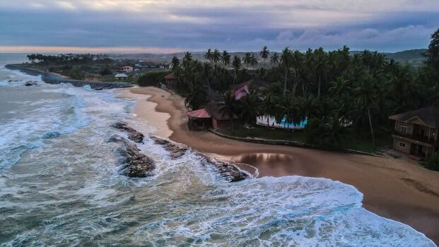 Aerial view of a tropical beach resort featuring lush palm trees a swimming pool and rocky breakwaters along the sandy coastline with crashing waves in Ghana. Aerial view of a tropical beach resort featuring lush palm trees a swimming pool and rocky breakwaters along the sandy coastline with crashing waves in Ghana.