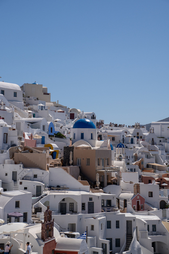 View of traditional white washed houses and blue domed churches on the volcanic cliffs with people walking along the pathways in Fira Greece. View of traditional white washed houses and blue domed churches on the volcanic cliffs with people walking along the pathways in Fira Greece.