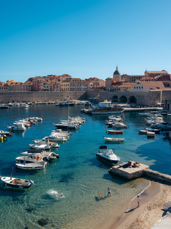 View of Dubrovnik Old Town harbor with small boats moored in turquoise water ancient stone walls and people on a small beach in Dubrovnik Dubrovnik Neretva County Croatia. View of Dubrovnik Old Town harbor with small boats moored in turquoise water ancient stone walls and people on a small beach in Dubrovnik Dubrovnik Neretva County Croatia.