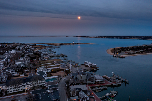 View of Martha s Vineyard and Edgartown harbor with a full moon reflecting on the water coastal houses and distant lighthouse at twilight Edgartown Massachusetts United States.