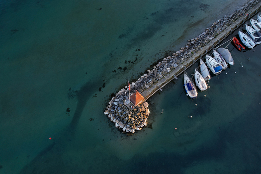 Aerial view of a stone pier with a small red roofed hut and sailboats docked in the clear turquoise waters of Lake Geneva Morges Vaud Switzerland. Aerial view of a stone pier with a small red roofed hut and sailboats docked in the clear turquoise waters of Lake Geneva Morges Vaud Switzerland.