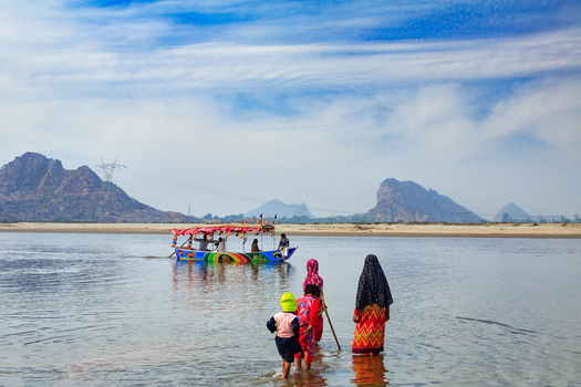 View of people wading in the shallow water of the Chenab River with a colorful boat in the background and mountains under a blue sky in Chiniot Punjab Pakistan.