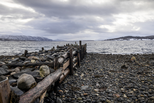View of a rustic wooden breakwater and rocky shoreline overlooking a calm fjord with snow capped mountains under a cloudy sky in Kvaloya Troms Norway. View of a rustic wooden breakwater and rocky shoreline overlooking a calm fjord with snow capped mountains under a cloudy sky in Kvaloya Troms Norway.
