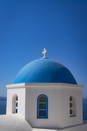 View of a traditional white church with a vibrant blue dome and a cross set against the deep blue Aegean Sea and a clear sky in Fira Greece.