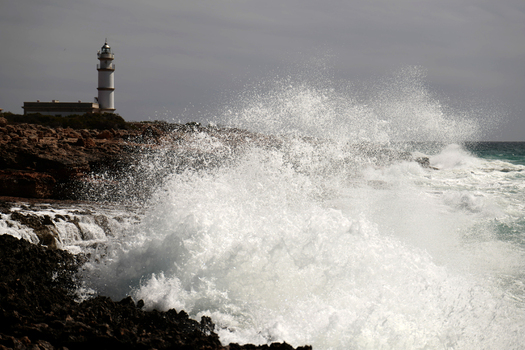 View of the lighthouse at Cap de Ses Salines with powerful waves crashing against the rocky coastline in Mallorca Balearic Islands Spain.