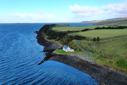 Aerial view of a solitary house situated on the rocky coastline overlooking the deep blue sea surrounded by green fields and rolling hills in Stein Scotland United Kingdom. Aerial view of a solitary house situated on the rocky coastline overlooking the deep blue sea surrounded by green fields and rolling hills in Stein Scotland United Kingdom.