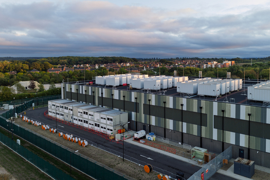 Aerial view of a large data center facility with modular cooling units on the roof fenced perimeter and surrounding suburban landscape in the United Kingdom. Aerial view of a large data center facility with modular cooling units on the roof fenced perimeter and surrounding suburban landscape in the United Kingdom.