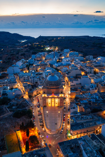 Aerial view of the Parish Church of the Assumption of the Blessed Virgin Mary into Heaven illuminated at dusk surrounded by the town of Mgarr Northern Region Malta.