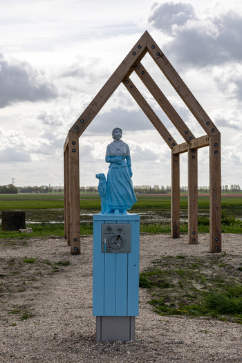 View of a blue statue of a woman and dog standing under a wooden house frame in the flat landscape of Schokland Flevoland Netherlands.