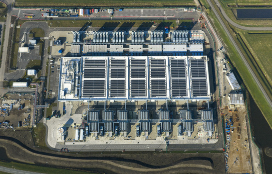 Aerial view of a digital data center facility featuring solar panels on the roof and cooling infrastructure surrounded by green fields Netherlands. Aerial view of a digital data center facility featuring solar panels on the roof and cooling infrastructure surrounded by green fields Netherlands.