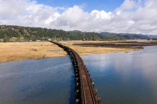 Aerial view of the Coos Bay Rail Line curving through marshland near Florence Oregon with calm water forested banks and tidal wetlands along the lower Siuslaw River.