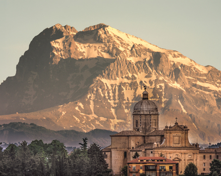 View of the historic cathedral with its prominent dome set against the backdrop of a snow capped mountain peak under soft light in Macerata Marche Italy. View of the historic cathedral with its prominent dome set against the backdrop of a snow capped mountain peak under soft light in Macerata Marche Italy.