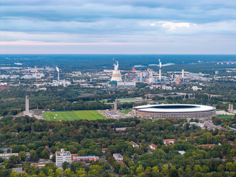 Aerial view of Olympiastadion Berlin surrounded by lush green trees urban infrastructure and a power plant in the distance under a cloudy sky in Berlin Berlin Germany.
