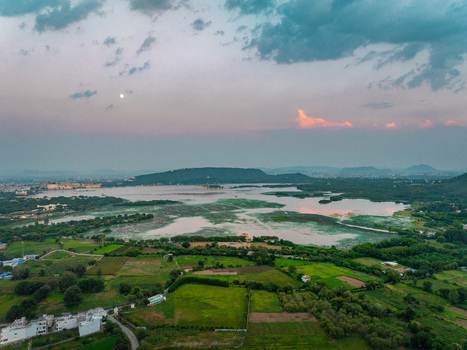 Aerial view of Lake Pichola surrounded by lush green fields historic buildings and the city skyline of Udaipur in Udaipur Rajasthan India.