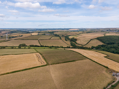 Aerial view of agricultural farmland featuring harvested fields hedgerows tracks and a wind turbine under a blue sky with clouds in Looe Cornwall United Kingdom. Aerial view of agricultural farmland featuring harvested fields hedgerows tracks and a wind turbine under a blue sky with clouds in Looe Cornwall United Kingdom.