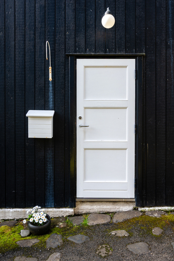 View of a white wooden door set against a black vertical timber wall with a matching white mailbox a small potted plant with white flowers and a mossy stone path in the Faroe Islands. View of a white wooden door set against a black vertical timber wall with a matching white mailbox a small potted plant with white flowers and a mossy stone path in the Faroe Islands.
