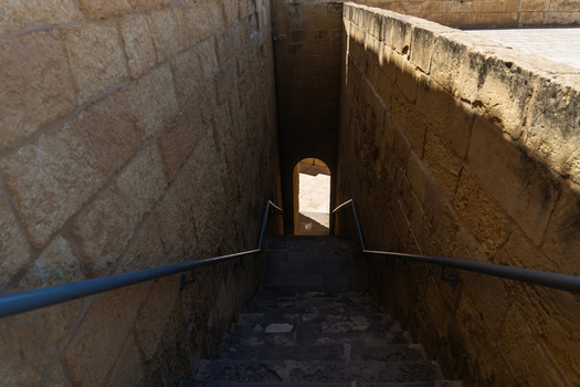 View of a narrow stone staircase descending between high walls toward an arched doorway in bright sunlight Zebbug Gozo Region Malta. View of a narrow stone staircase descending between high walls toward an arched doorway in bright sunlight Zebbug Gozo Region Malta.