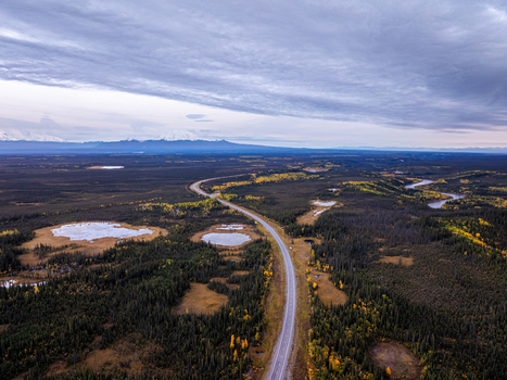 Aerial view of the highway winding through the vast boreal forest and wetlands near Paxson with distant snow capped mountains under a cloudy sky in Paxson Alaska United States. Aerial view of the highway winding through the vast boreal forest and wetlands near Paxson with distant snow capped mountains under a cloudy sky in Paxson Alaska United States.