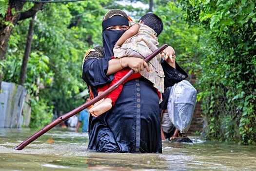 Banshkhali Upazila Chittagong Division Bangladesh 10 July 2026 View of a woman in a niqab carrying a child through floodwaters while using a wooden stick for balance. Banshkhali Upazila Chittagong Division Bangladesh 10 July 2026 View of a woman in a niqab carrying a child through floodwaters while using a wooden stick for balance.
