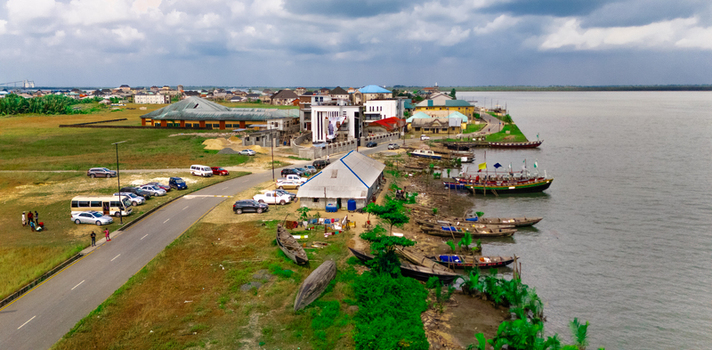 Aerial view of the coastal town of Minima with traditional wooden boats docked along the riverbank nearby buildings a road with parked cars and lush green vegetation in Minima Rivers Nigeria.