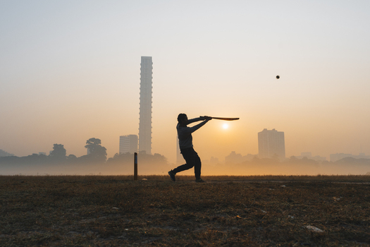 View of a lone cricketer silhouetted against the warm glow of the rising sun playing a shot with cityscape in the misty background Kolkata West Bengal India. View of a lone cricketer silhouetted against the warm glow of the rising sun playing a shot with cityscape in the misty background Kolkata West Bengal India.