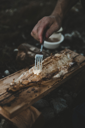 View of a person placing butter on a piece of plank grilled fish on a wooden board in Laane County Estonia. View of a person placing butter on a piece of plank grilled fish on a wooden board in Laane County Estonia.