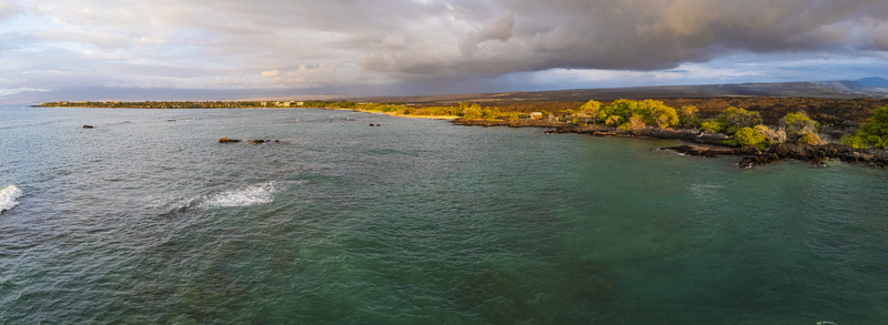 Aerial view of Waikoloa Beach featuring turquoise ocean waters dark volcanic rock formations and lush green vegetation under a dramatic cloudy sky at sunset Waikoloa Village Hawaii United States.
