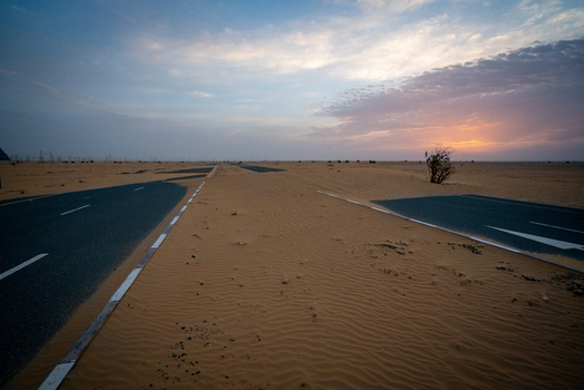 View of an asphalt road partially covered by shifting sand dunes in the desert under a soft sunset sky Dubai Dubai United Arab Emirates. View of an asphalt road partially covered by shifting sand dunes in the desert under a soft sunset sky Dubai Dubai United Arab Emirates.