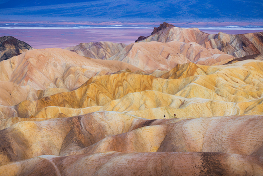View of the golden badlands of Death Valley National Park with two hikers walking along a ridge under a clear sky in the United States.