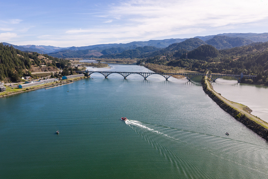 Aerial view of the Rogue River and Isaac Lee Patterson Bridge with salmon fishing boats surrounded by lush hills under a clear blue sky in Gold Beach Oregon United States.
