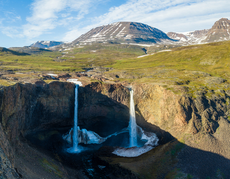 Aerial view of two waterfalls cascading into a pool with snow and ice surrounded by rocky terrain and mountains in Greenland. Aerial view of two waterfalls cascading into a pool with snow and ice surrounded by rocky terrain and mountains in Greenland.